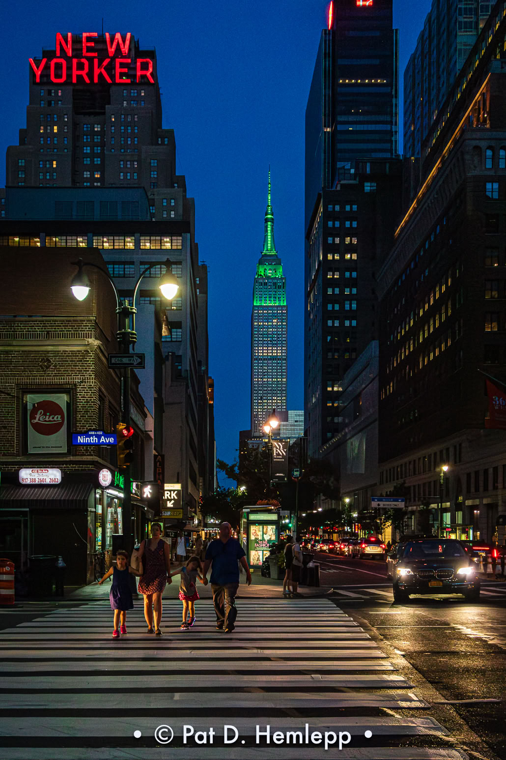The Empire State Building, illuminated in green, stands behind pedestrians on 34th Street as they cross Ninth Avenue in New York City.